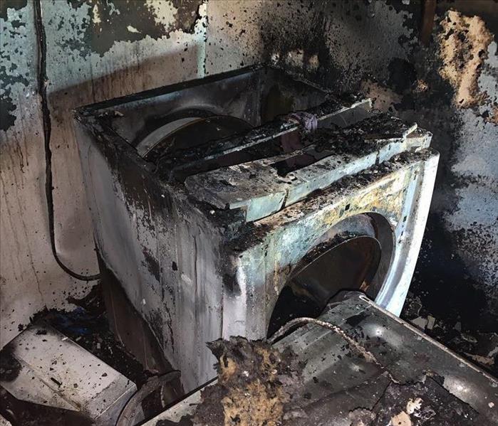A heavily fire-damaged laundry room with particular emphasis on damage to an electric dryer and adjacent washer