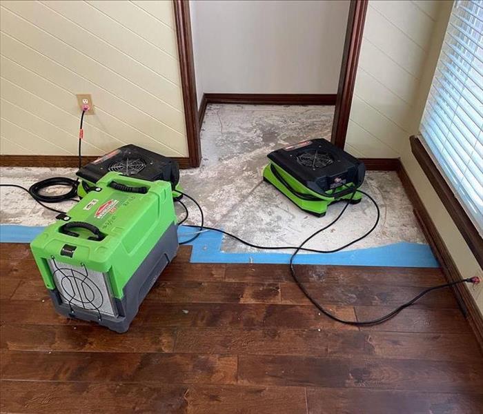 Two SERVPRO green air movers and a dehumidifier on a partially demolished wood floor with a visibly tarnished subfloor